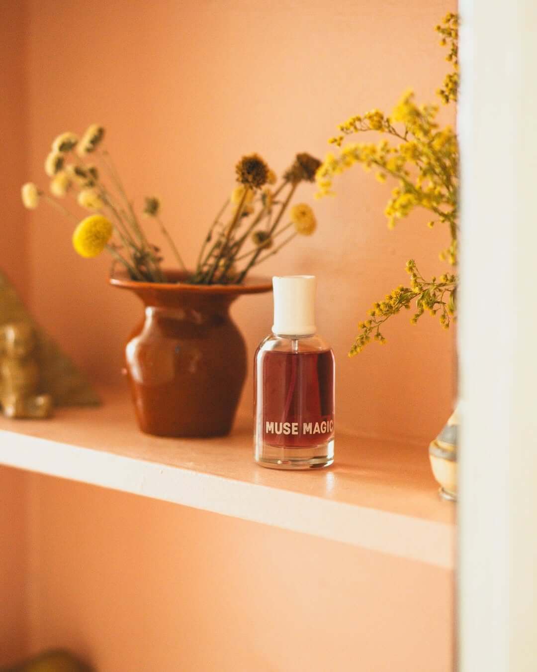 Bottle of Muse Magic Eau de Parfum on a shelf with decorative vases and flowers in the background.